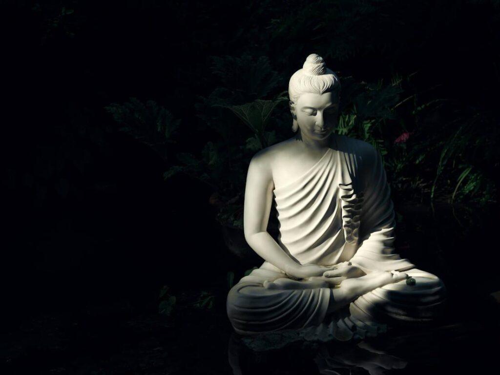 Vit staty av Buddha i sten med växter bakom.